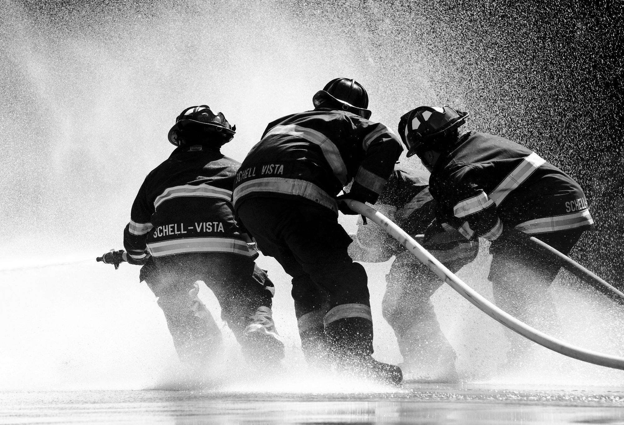ICRA - A black and white image captures three firefighters in protective gear, marked "SCHELL-VISTA," as they work with urgency to control a high-pressure hose, water spraying around them. The scene, reminiscent of an ICRA training exercise, powerfully conveys teamwork and determination.