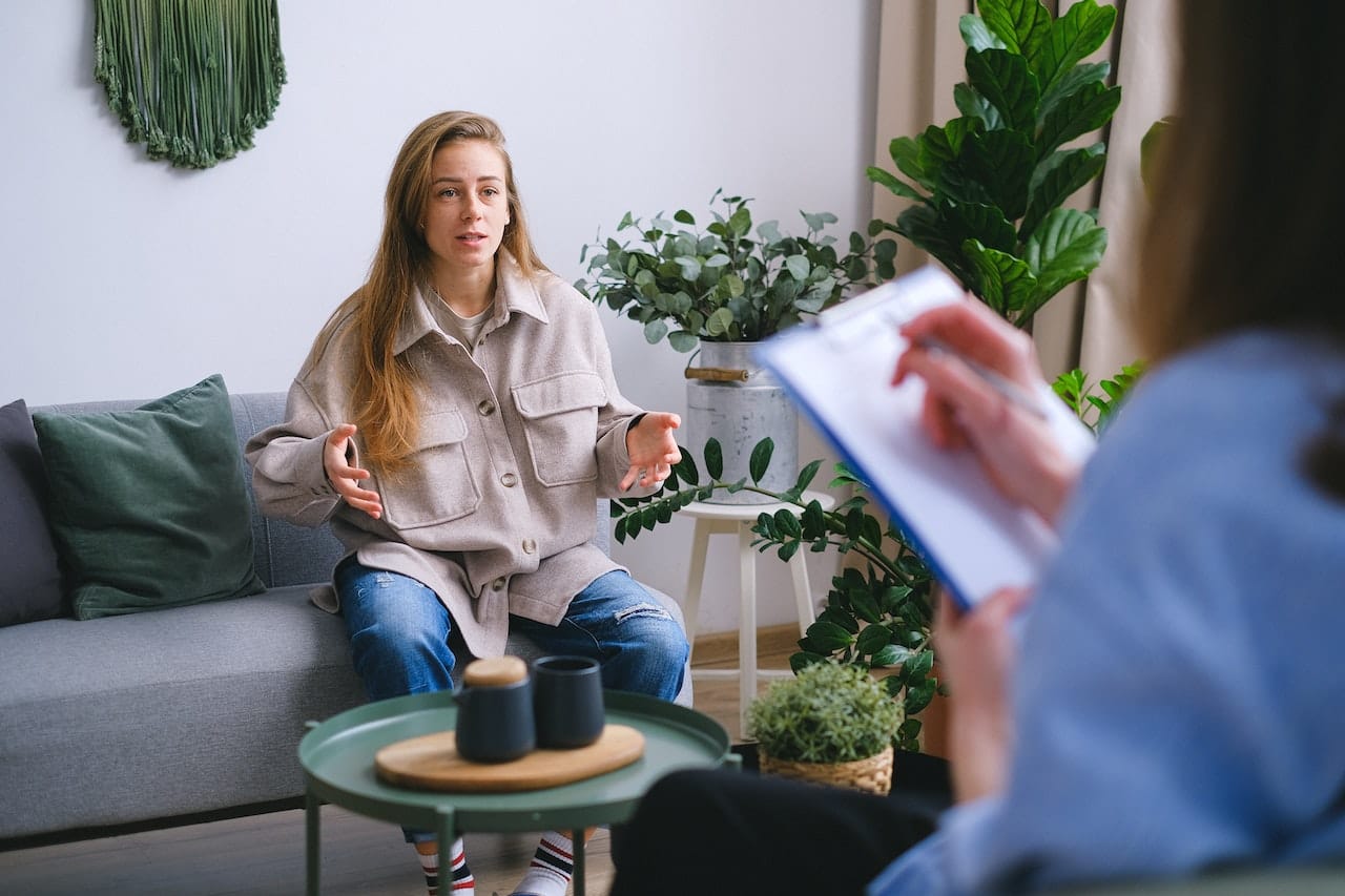 ICRA - A woman sits on a couch, gesturing energetically during a conversation about ICRA. In the foreground, a person holds a clipboard, attentively taking notes. The room is cozy, adorned with lush plants and soft furnishings.