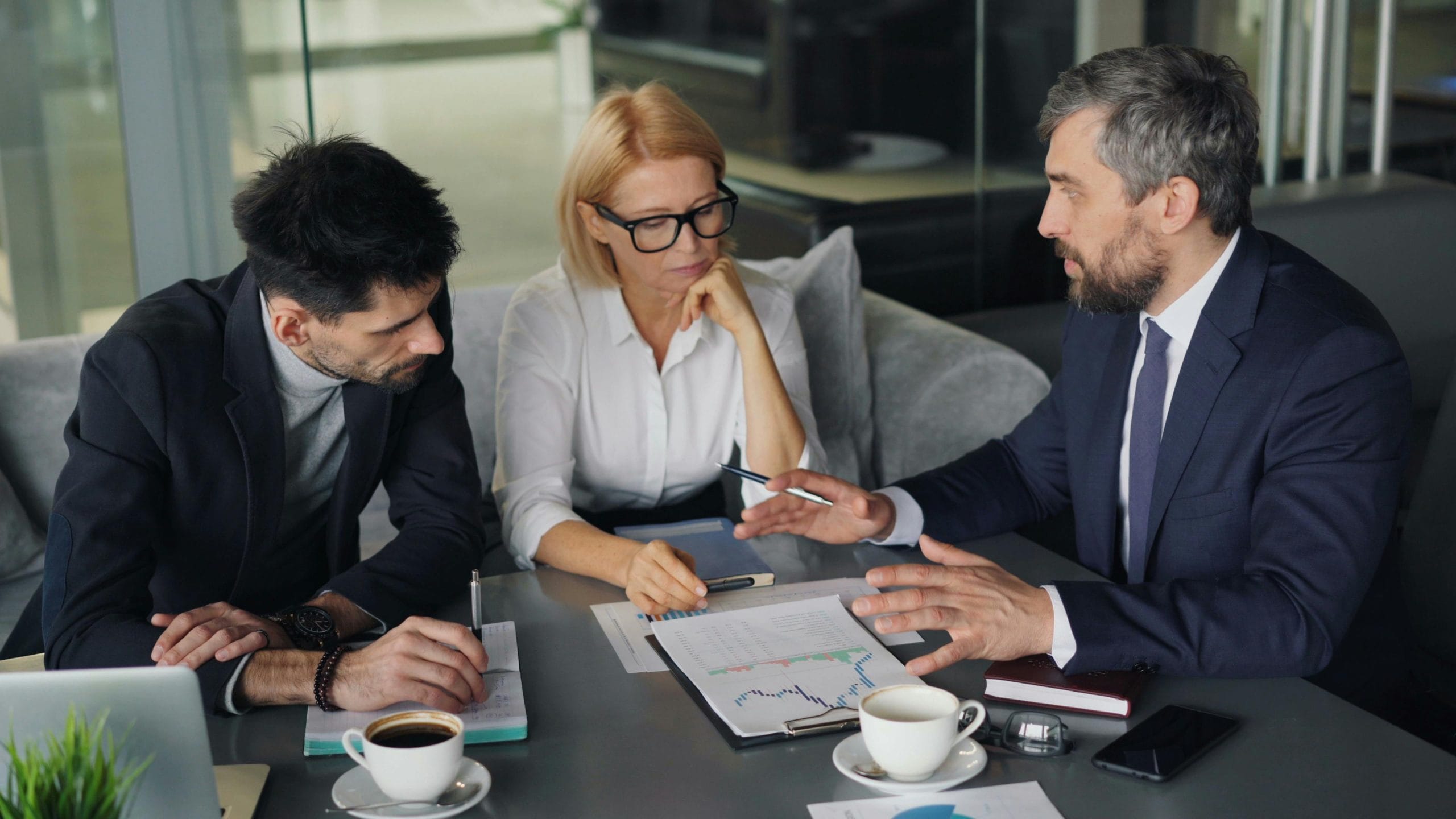 ICRA - Three business professionals in a meeting room are engaged in discussion over financial documents, referencing the latest ICRA ratings. A man in a suit speaks, while a woman with glasses and another man listen attentively. Coffee cups and papers are scattered on the table.