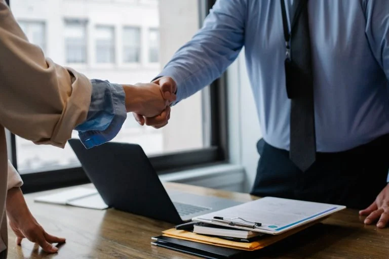 ICRA - Two people are shaking hands over a wooden desk with a laptop, a clipboard, and documents, showcasing the professional aura common in ICRA meetings. Dressed in business attire, the scene hints at a pivotal business agreement or meeting near a large window.