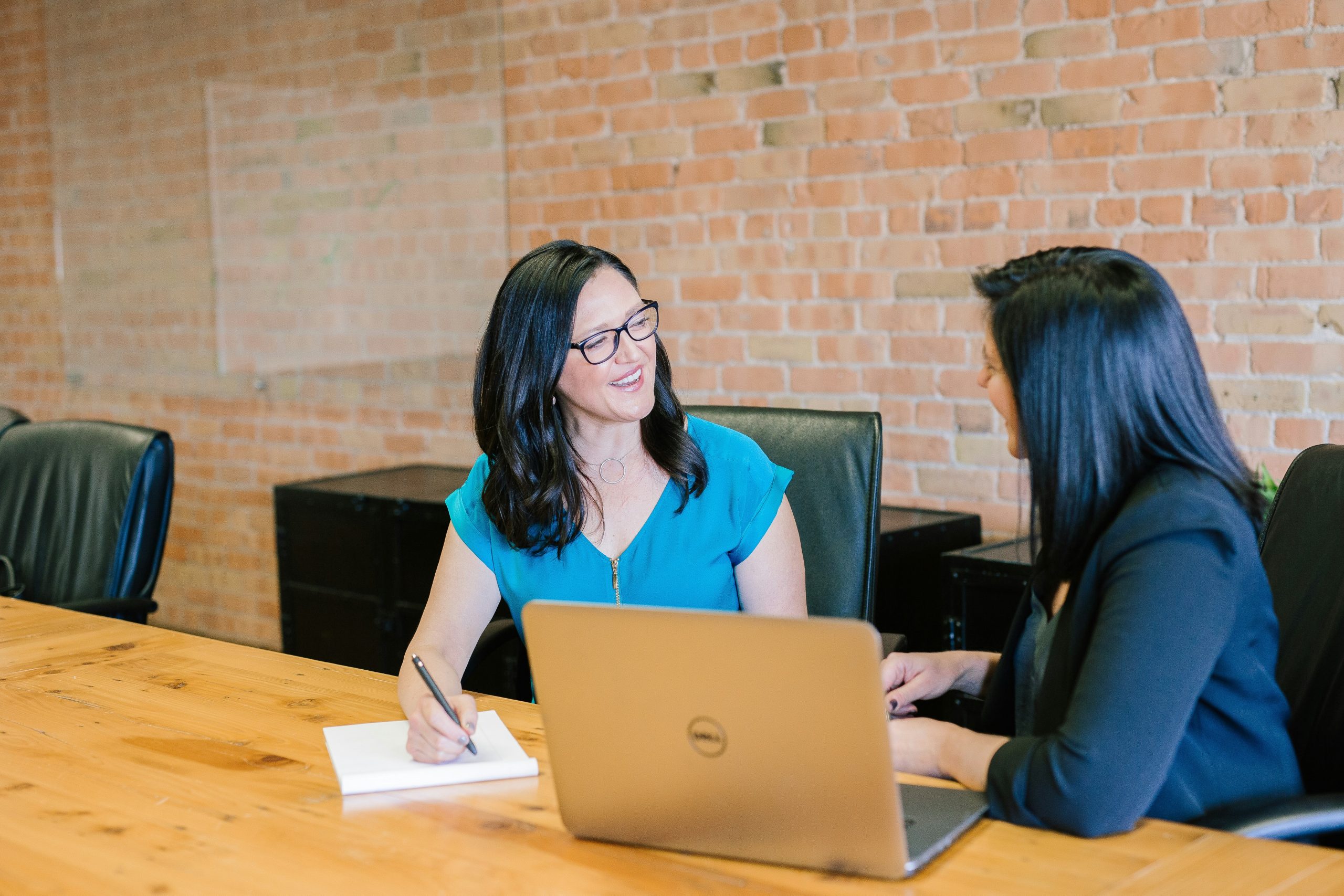 ICRA - Two women are sitting at a wooden table in an office setting, engaged in conversation about the upcoming ICRA conference. One woman is writing on a notepad, while the other listens intently with a laptop open in front of her. A brick wall serves as the backdrop to their discussion.